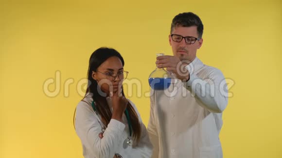 多种族的男人和女人穿着实验室外套在黄色背景上用实验液看着瓶子视频的预览图