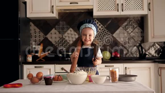 一个戴着帽子和围裙的小女孩用木勺吃生饼干面团视频的预览图