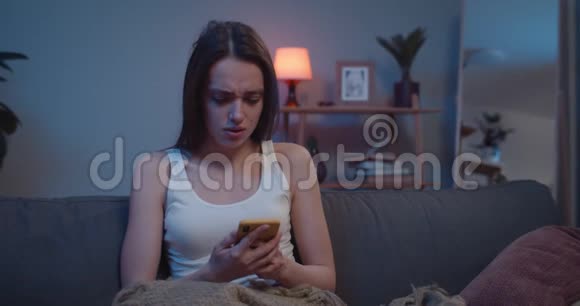 不高兴的年轻女孩在家里坐在沙发上用智能手机获取床上新闻女千禧一代看上去很担心视频的预览图