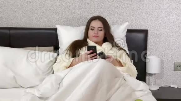 躺在床上用智能手机收发信息切换电视频道的特大号年轻女性视频的预览图