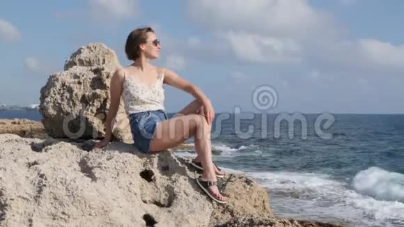 年轻性感的白种人女人美丽的身体坐在海边的岩石上看着大海迷人的女性享受阳光视频的预览图