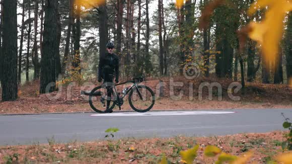 迷人的年轻骑自行车的人穿着自行车衣服黑色头盔和运动太阳镜在秋天的森林里做的动作快乐英俊视频的预览图