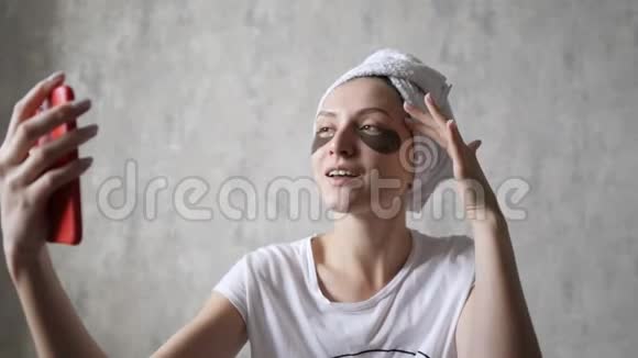 女孩正在家里闲逛一个年轻的女人眼睛下面贴着补丁一条毛巾看着手机的摄像头视频的预览图