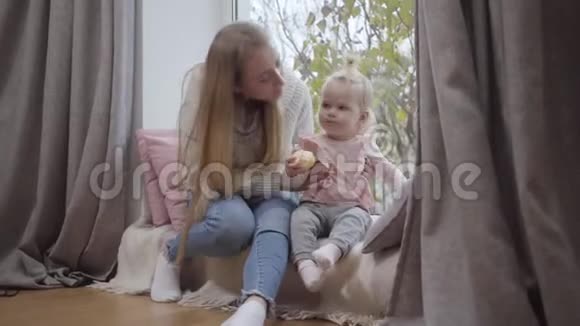 幸福的白种人年轻母亲的特写亲吻她可爱的宝贝女儿的脸颊和微笑小金发姑娘视频的预览图