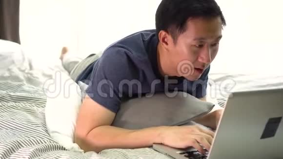 年轻微笑的亚洲男人穿着休闲服装躺在床上用笔记本电脑在家工作的男性自由职业者视频的预览图