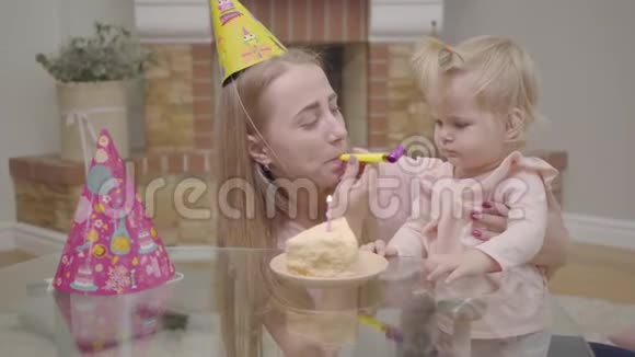 年轻的白种人母亲戴着派对帽小可爱的女婴玩着派对哨子一块蛋糕和一支蜡烛视频的预览图