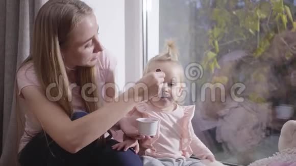 白种人年轻女人坐在低矮的窗台上喂小金发女孩的肖像漂亮的妈妈照顾视频的预览图