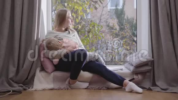 年轻快乐的高加索女人坐在低矮的窗台上双手抱着女儿的肖像微笑的母亲视频的预览图