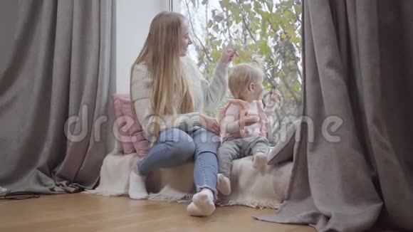 快乐的白人母女坐在窗台上透过窗户向外看可爱的金发女婴视频的预览图
