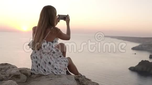 女人在日落时欣赏海景并在手机上拍照坐在悬崖上的长发女孩视频的预览图