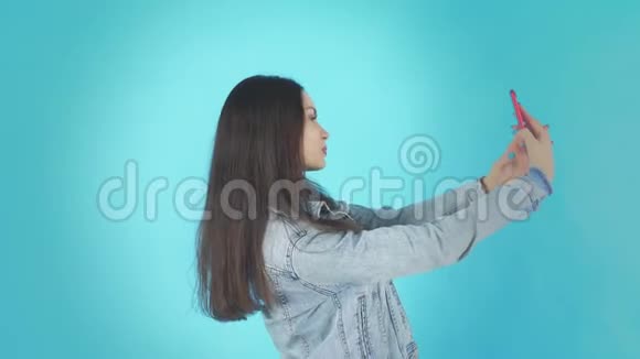 戴尼姆夹克中微笑快乐的女人在蓝底智能手机上做自我视频的预览图