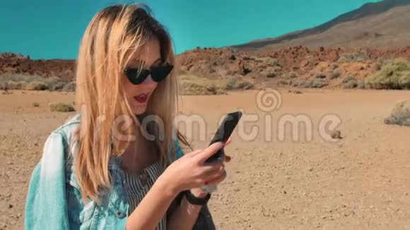 穿牛仔夹克戴太阳镜的女人用手机发信息火山背景下的沙漠视频的预览图