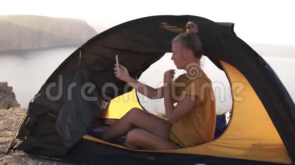 穿着黄色T恤的苗条金发女孩坐在露营帐篷旁边的地上一边看一边梳头视频的预览图