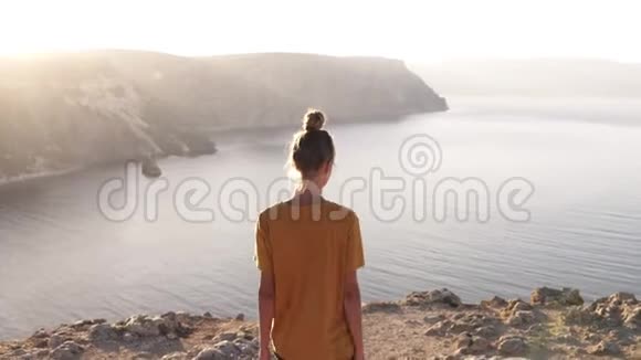 一个穿着黄色T恤和短裤的苗条女孩走在山的边缘在下面欣赏风景视频的预览图
