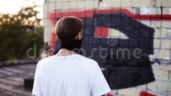 一个穿着白色T恤和黑色围巾的年轻人带着涂鸦走向一堵墙还在继续视频的预览图