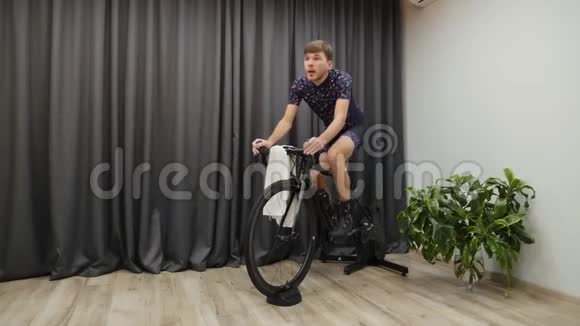 室内自行车教练的专业自行车训练男运动员在家骑自行车出汗手拿毛巾和视频的预览图