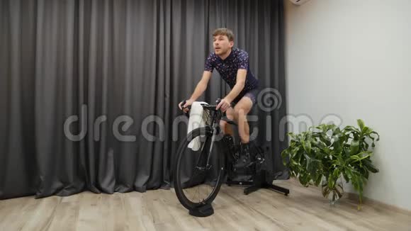 人累后骑自行车硬练自行车智能培训师男的在室内骑自行车完成了踏板年轻运动员在做视频的预览图