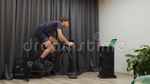 年轻的运动员从马鞍上骑自行车并在家庭智能自行车教练上密集地踏板室内用重型齿轮踏板Cycli视频的预览图