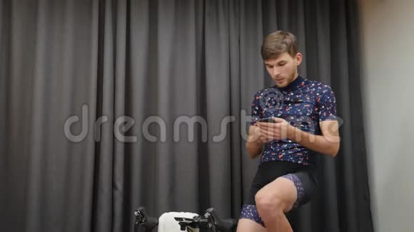 室内自行车的概念男性看电话和脚踏自行车智能教练一个在家骑自行车锻炼身体的人视频的预览图