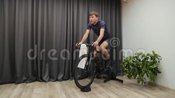 男性自行车手在家训练自行车智能教练手拿毛巾和擦拭男子骑自行车准备骑自行车视频的预览图