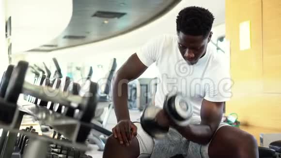 年轻的非裔美国人坐着举起一个哑铃靠近健身房的架子男性体重训练者做视频的预览图