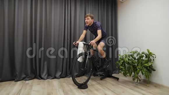 骑自行车在智能教练室内锻炼男性出汗时循环训练解开球衣和拿毛巾骑自行车虚拟视频的预览图