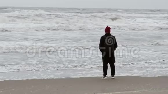 秋天戴着红帽子的年轻潮人从沙滩上的海浪中退回来视频的预览图