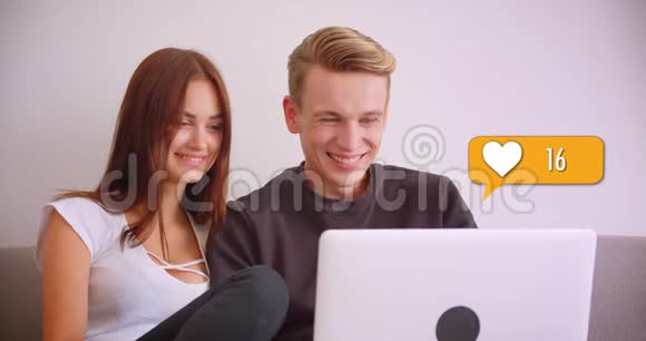 可爱快乐的白种人夫妇在社交媒体上使用笔记本电脑坐在室内沙发上获得喜欢的特写肖像视频的预览图