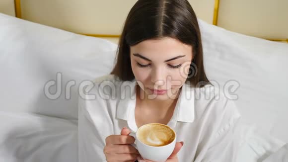 年轻漂亮自信的女人坐在白色床上捧着咖啡杯微笑着看着视频的预览图