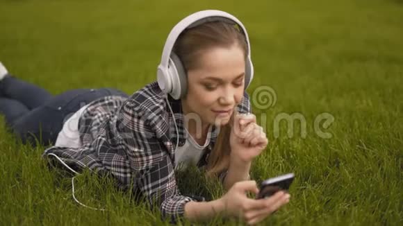 在网上搜索一个年轻的女人戴着大耳机躺在草地上看着视频视频的预览图