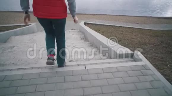 阴天时穿着无袖红色夹克的女孩沿着台阶走到湖边视频的预览图