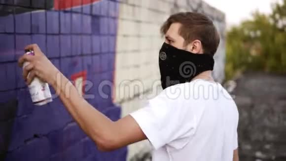 一个年轻的白种人的侧视图他穿着白色T恤脸上戴着黑色围巾用气雾剂涂上蓝色颜料视频的预览图