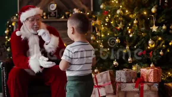 小男孩来到微笑的圣诞老人身边圣诞老人坐在圣诞树旁边视频的预览图