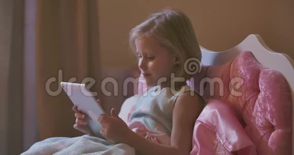 漂亮的金发白种人女孩躺在粉红色的床上用平板电脑的肖像现代儿童在看动画片之前视频的预览图