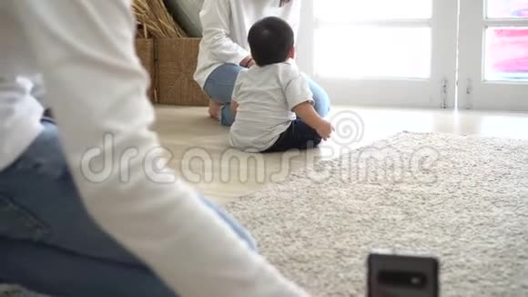 家长用智能手机拍摄可爱的亚洲宝宝在家里柔软的白地毯上爬行的视频视频的预览图