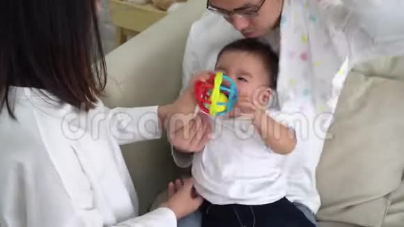 年轻的亚洲父亲坐在地板上抱着婴儿而母亲则拿着婴儿衣服一起照顾孩子视频的预览图