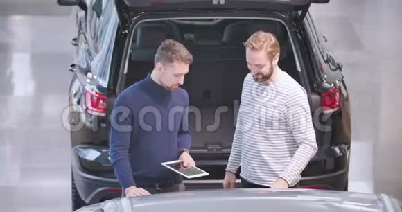 两个白人男子站在汽车经销商和交谈男性汽车经销商与平板电脑解释规格视频的预览图