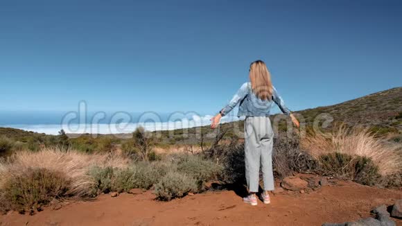 穿着牛仔夹克和条纹裤子的金发女人站在云层上方的一座高山上举起双手这就是视频的预览图