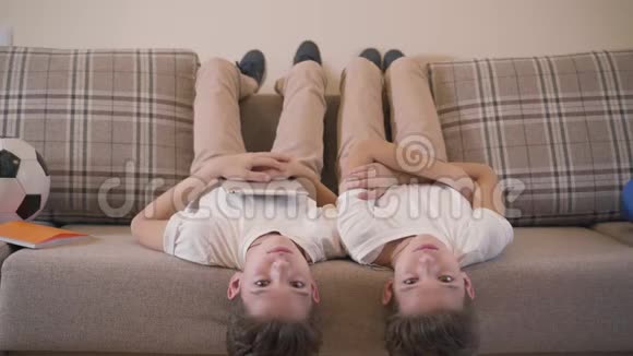 两个高加索双胞胎兄弟倒躺在沙发上看着摄像机笑着快乐的兄弟姐妹视频的预览图