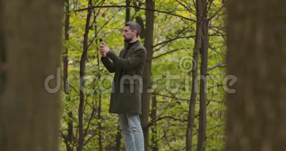 微笑着一个白发苍苍的白人男子站在秋天森林的树木之间在他的智能手机上拍照视频的预览图