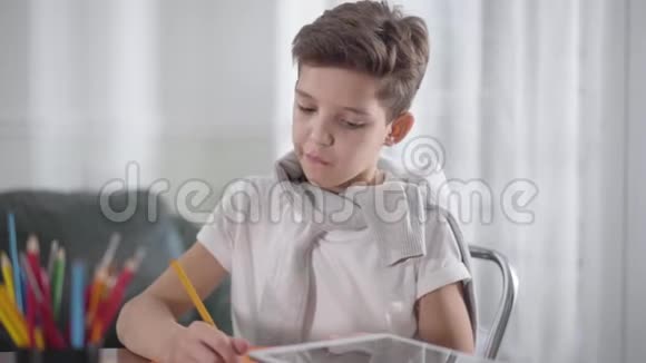 集中白人男孩坐在桌子旁在笔记本上画画或写作的肖像小学生做作业视频的预览图