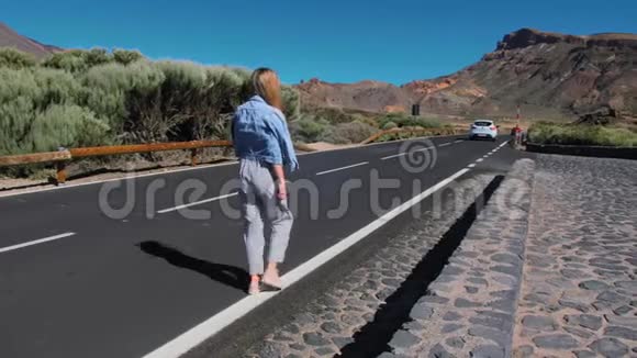 穿着牛仔夹克和条纹裤子的女孩沿着高地的一条沥青路走到远处视频的预览图