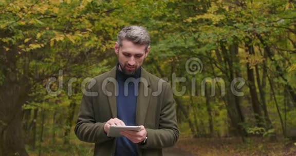 英俊的高加索游客站在秋天的森林里在他的平板电脑上打字微笑着自信的男人分享视频的预览图