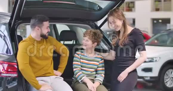 年轻的白种人父亲和母亲微笑着拥抱他们的儿子幸福的家庭在展厅选择新汽车视频的预览图