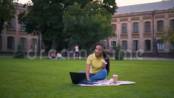 更远的距离专心致志的女孩在公园里坐在草坪上女孩在笔记本电脑上工作吃三明治4K视频的预览图
