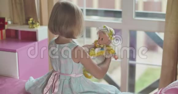 小白种人金发女孩坐在粉红色的床上玩娃娃穿着漂亮蓝色衣服的孩子视频的预览图