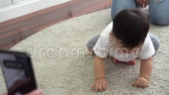 家长用智能手机拍摄可爱的亚洲宝宝在家里柔软的白地毯上爬行的视频视频的预览图