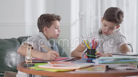 两个高加索双胞胎兄弟坐在桌子旁学习一个男孩试图作弊他的兄弟关闭了他的锻炼视频的预览图