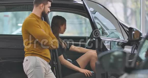 坐在汽车沙龙里和站在车旁的男朋友或丈夫聊天的美丽白人女孩视频的预览图
