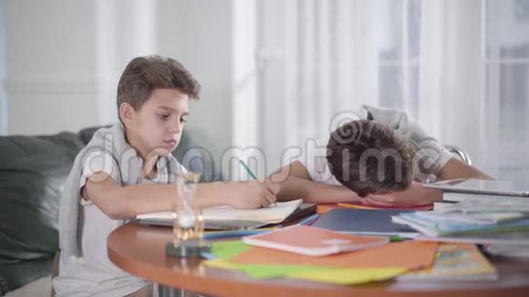 聪明的白人男孩做家庭作业的肖像他的兄弟睡在桌子上孩子醒来时他的兄弟姐妹视频的预览图